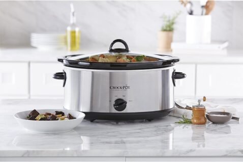 A stainless steel Crock-Pot slow cooker on a kitchen counter, filled with vegetables and stew, with a serving bowl of food placed beside it.