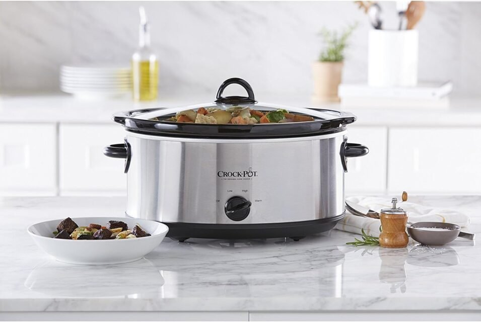 A stainless steel Crock-Pot slow cooker on a kitchen counter, filled with vegetables and stew, with a serving bowl of food placed beside it.