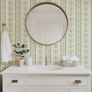 White bathroom vanity with gold accents, floral wallpaper, round mirror, and neatly arranged towels and decor.