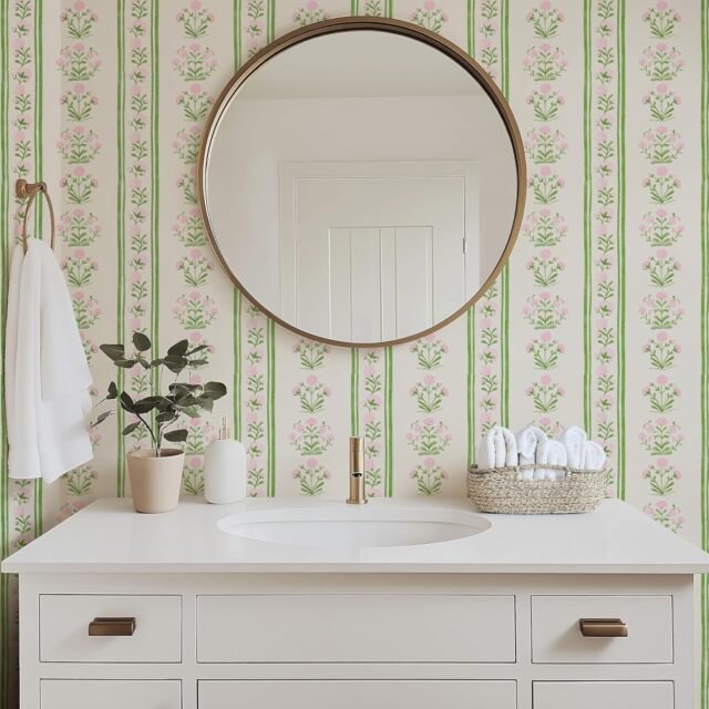 White bathroom vanity with gold accents, floral wallpaper, round mirror, and neatly arranged towels and decor.
