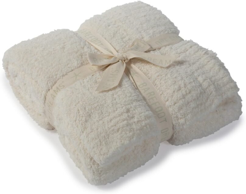 A cream-colored fluffy blanket neatly folded and tied with a beige ribbon labeled “BAREFOOT DREAMS.”